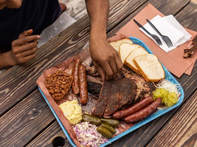 Plastic tray covered in butcher paper topped with slices of different barbecue meats and traditional sides.