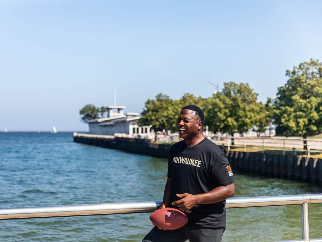 A man, LeRoy Butler, holding a football and smiling while leaning on a metal railing by a waterfront. Trees line the pathway behind him, and a building extends into the water in the background. The weather is clear and sunny.
