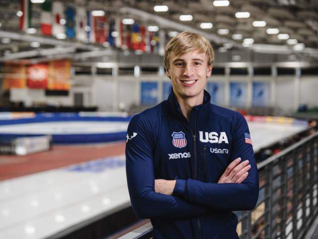 A person wearing a navy USA team jacket stands smiling on an indoor ice rink, with the rink blurred in the background. The jacket displays several sponsor logos and a small American flag patch.