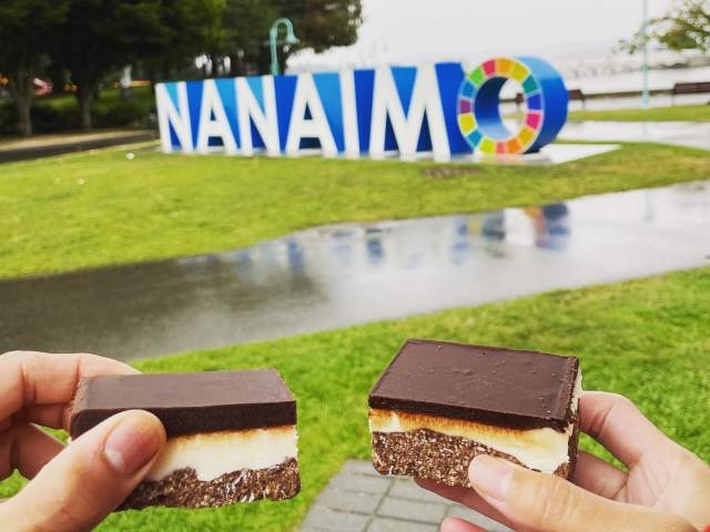 Two people cheering Nanaimo bars in front of the Nanaimo sign.