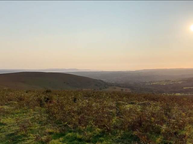 A rural Welsh landscape with the sea in the distance and a low sun in the sky.