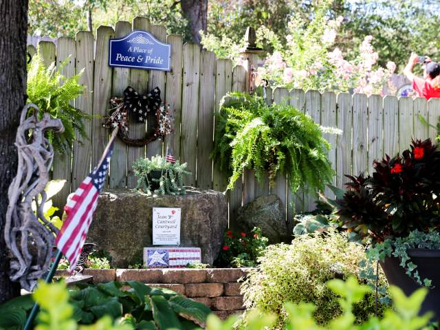 A beautifully landscaped memorial display inside the Branson Veterans Memorial Garden in Branson, Missouri. The focal point is a weathered wooden fence adorned with a blue arched sign that reads "A Place of Peace & Pride" in white lettering. Below the sign hangs a patriotic wreath with a black and white star-patterned bow. A large natural boulder serves as a pedestal for a small potted plant, a white dedication plaque, and a painted patriotic brick. An American flag on a small staff projects forward from the left side of the display. The garden is lush with greenery, including vibrant green ferns hanging on the fence, leafy hostas, a stone-brick border wall, and a large dark planter filled with red flowers and trailing vines in the right foreground. In the blurry upper-right background, a visitor wearing a red shirt is seen taking a photograph behind the fence line under sunny, dappled light.