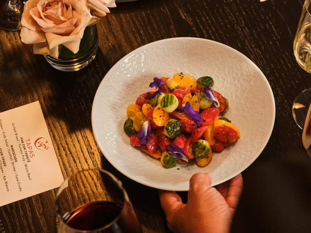 A dining table at Capa features a colorful tomato-based dish alongside a plate of thinly sliced cured ham, bread, wine glasses, menus, and floral accents. The image highlights the restaurant's Spanish-inspired cuisine and elevated food presentation within a fine dining setting.