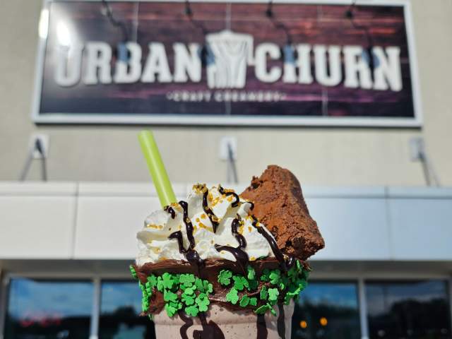 A person holding up a milkshake with whipped cream, a brown, and chocolate sauce in front of a sign that reads Urban Churn
