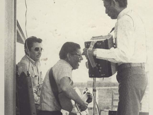 Historic photo of a small conjunto band including a guitar and an accordion player next to a building in the Montopolis neighborhood