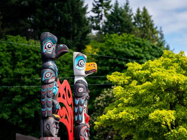 Two totem poles facing to the right with trees behind them.