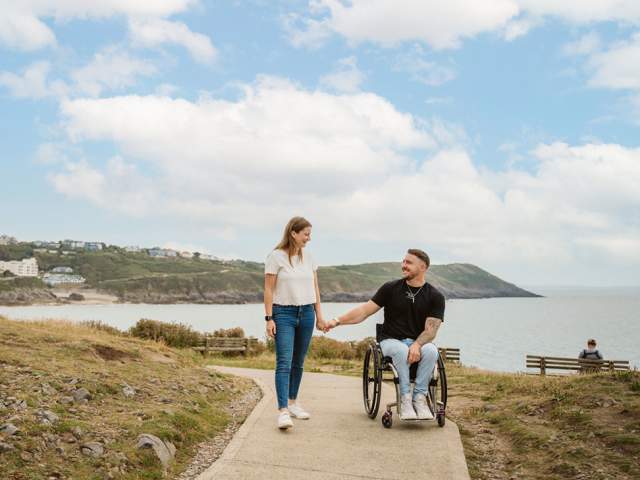 A man in a wheelchair holding hands with a standing woman on a section of the coast path with the sea in the background.