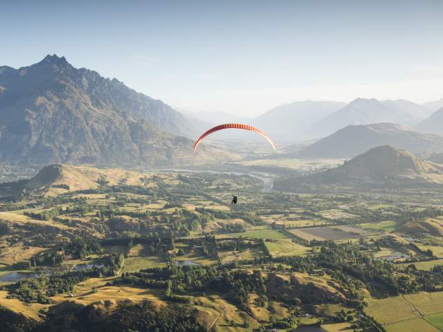 Person paragliding over a green valley with the Remarkables mountain range in the background