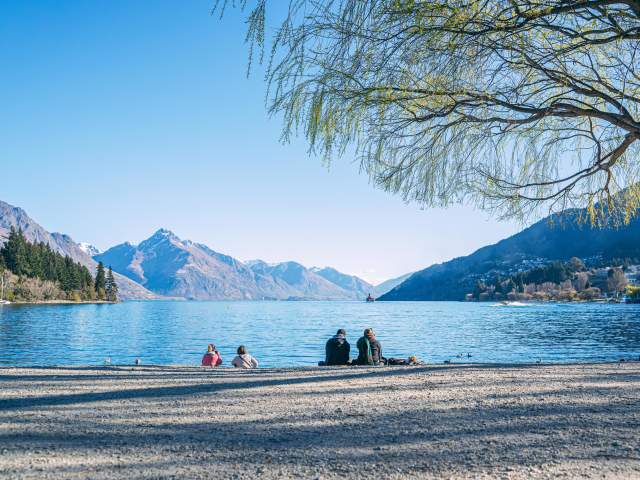 People sitting on Queenstown beach in spring with lake views and mountains in the background