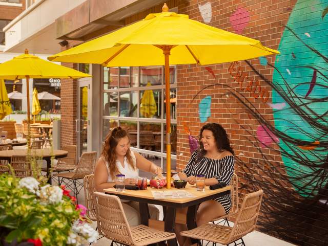 two women eating at an outdoor table at a restaurant sitting under a yellow umbrella with a mural on the wall behind them