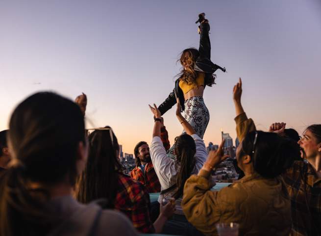 A woman dances with a crowd on a rooftop, overlooking a city skyline.