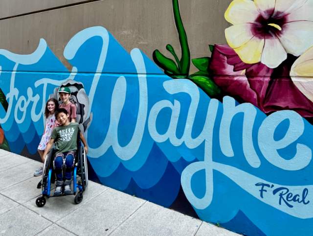 Two boys, one using a wheelchair, and a girl pose in front of a blue Fort Wayne script mural