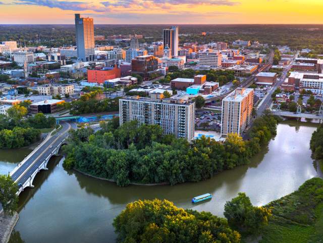 Drone shot of downtown Fort Wayne, Indiana at sunset with the confluence of the St Marys and St Joseph Rivers in the foreground and the skyline in the background. The Sweet Breeze canal boat is sailing on the St Marys River.