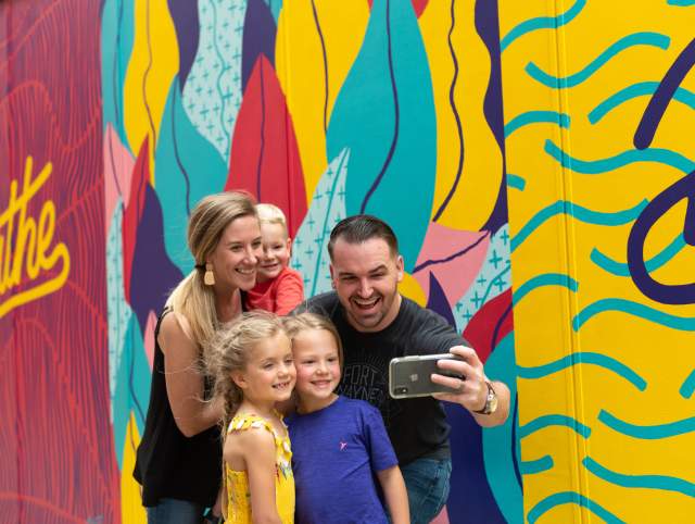 A family taking a cheerful selfie in front of the colorful 'Breathe' mural in downtown Fort Wayne.