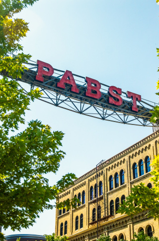 Historic Pabst Brewery buildings framed by leafy green trees, with the iconic red PABST sign spanning overhead against a bright blue sky.