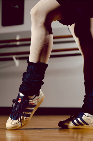 Close-up of a dancer’s legs and feet in sneakers and leg warmers, poised on a wooden studio floor near a ballet barre.