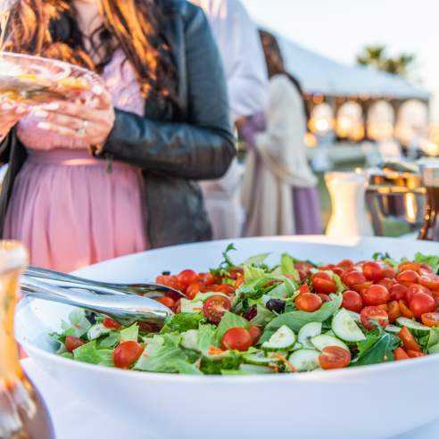 A large salad sits on a table while people wait in life for a buffet.