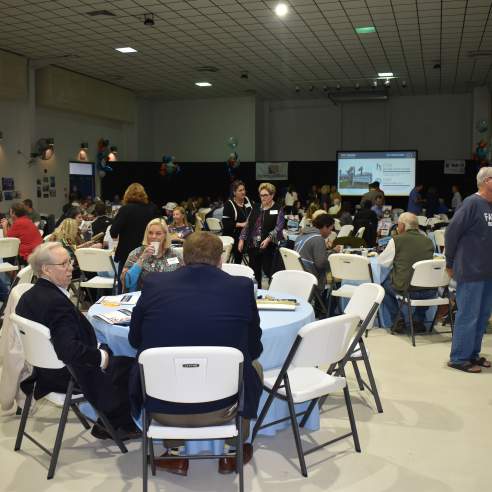 A room full of round tables with people sitting or standing around them.