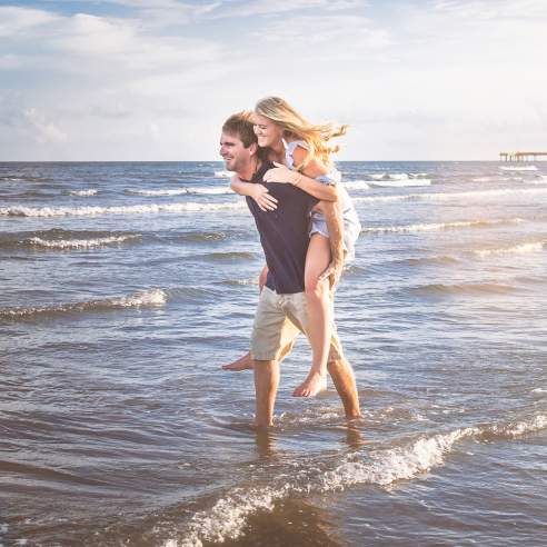 A man gives a woman a piggyback ride on the beach with the pier in the background in Port Aransas, TX