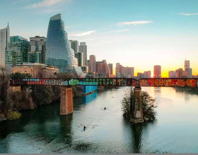 Kayakers paddling under a bridge with grafitti on Lady Bird Lake while the sun sets against Downtown Austin buildings.