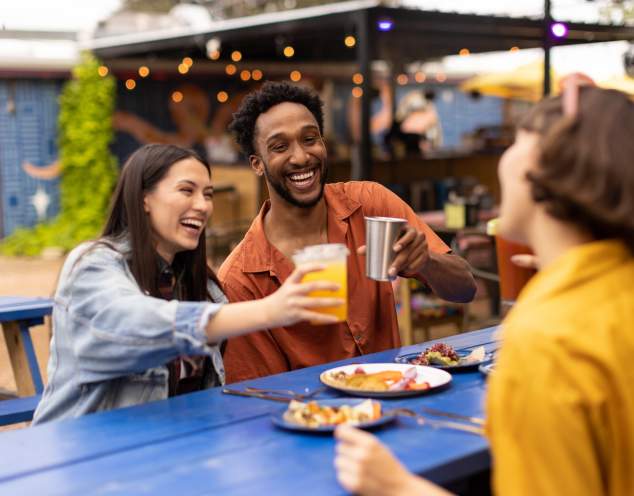 Two women and a man cheering drinks over plates of tacos.