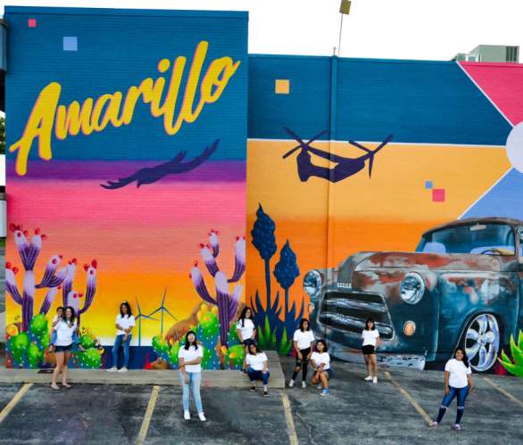 high school kids from the Blank Spaces Mural Group standing in front of a colorful mural they painted that shows a prairie dog driving a truck in from of a sunset with a Bell Boeing V-22 Osprey Fleet Surpasses 500,000 Flight Hours - Bell (news)Bell Boeing V-22 Osprey in the background