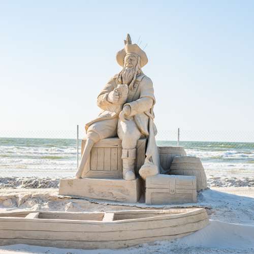 Sand sculpture of a pirate drinking out of a cup and sitting on barrels. In front of him is a rowboat made of sand