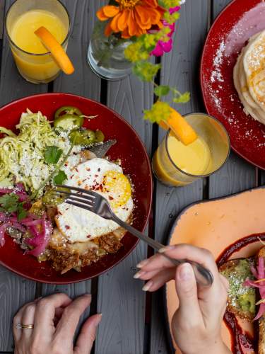 An overhead view of a table full of bright colored plates with various breakfast dishes and glasses of orange juice being served.