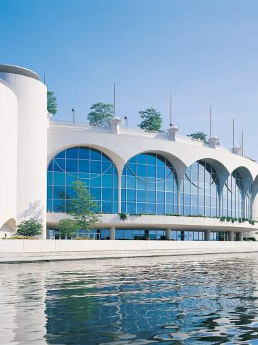 A view of Monona Terrace from Lake Monona. The sky is clear and sunny and a reflection of the building at its larger arched windows can be seen in the lake water.