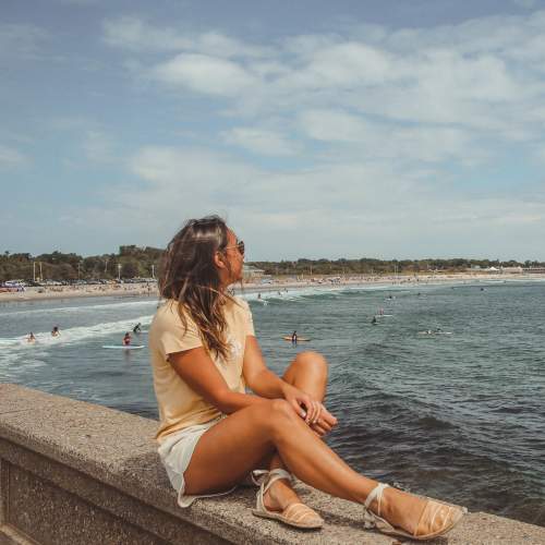 Woman sitting on a stone ledge by ocean watching surfers and beach under cloudy sky.