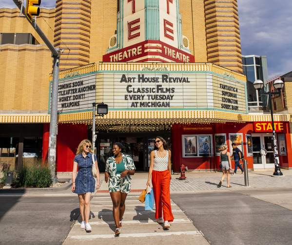 Three women in colorful clothing walking in the cross walk in front of the State Theatre