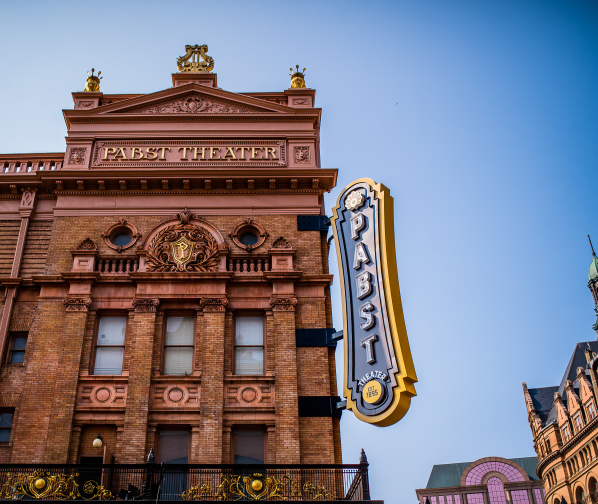The exterior of the historic Pabst Theater in Milwaukee, featuring its ornate brick façade, gold architectural details, and large vertical “Pabst Theater” marquee sign against a clear blue sky.