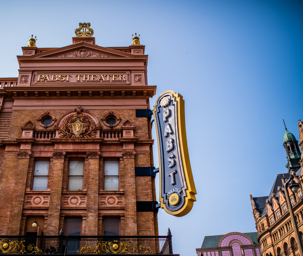 The exterior of the historic Pabst Theater in Milwaukee, featuring its ornate brick façade, gold architectural details, and large vertical “Pabst Theater” marquee sign against a clear blue sky.