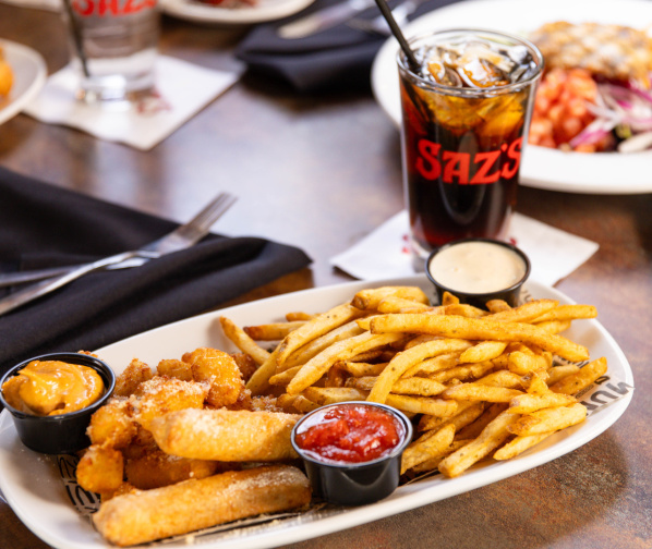 A close-up of a restaurant table featuring a platter of seasoned fries, mozzarella sticks, and dipping sauces. Behind it sits a glass of soda with the restaurant’s logo, and additional plates of food and black napkins are visible in the background.