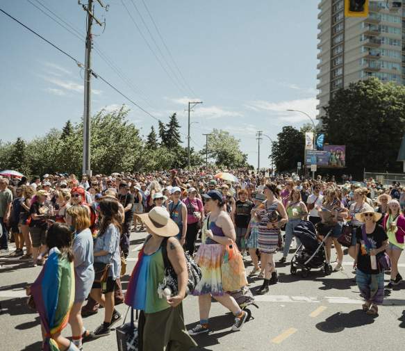 Hundreds of people take part in a Pride parade, filling a sunny Nanaimo street with rainbow colours, flags, and a celebratory atmosphere.