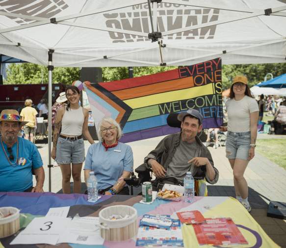 Five people smile under a tent at a community Pride event, seated at an information table with a rainbow ‘Everyone is welcome here’ flag displayed behind them.