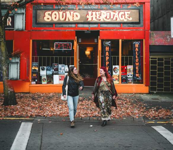 Two people walking past a red storefront with autumn leaves on the ground.