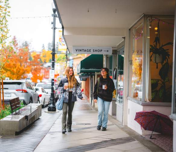 Two people stroll past shops on a rainy autumn day in downtown Nanaimo, holding coffee as colourful fall trees line the street.