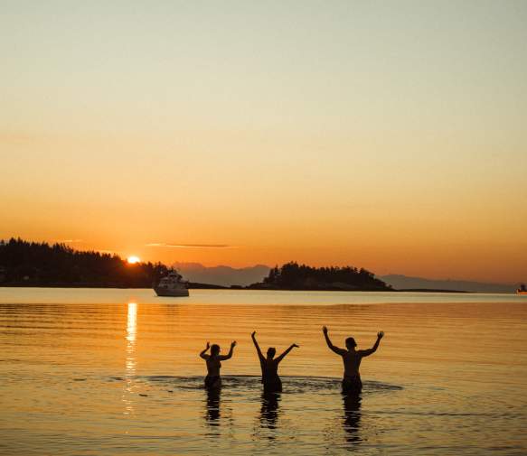 Three people standing in calm ocean water at sunset with arms raised.