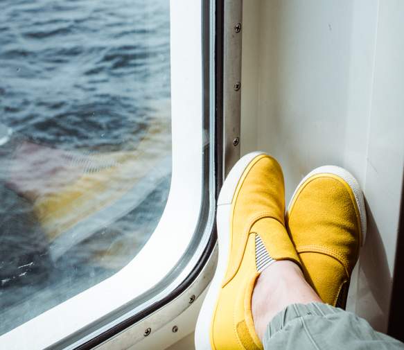 Person wearing yellow slip-on shoes resting their feet by a ferry window overlooking the ocean.