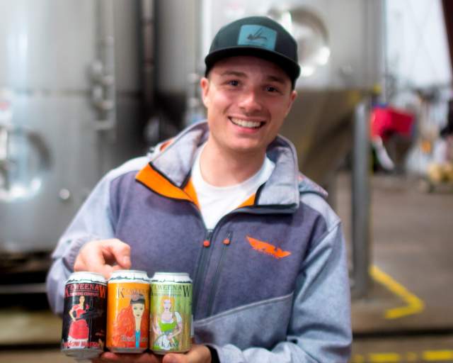 a person smiles while holding a six pack of beer, tall brewery vats behind them