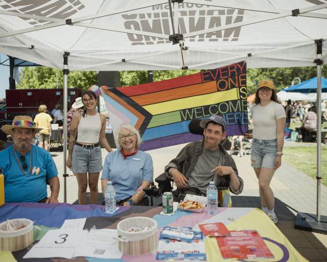 Five people smile under a tent at a community Pride event, seated at an information table with a rainbow ‘Everyone is welcome here’ flag displayed behind them.