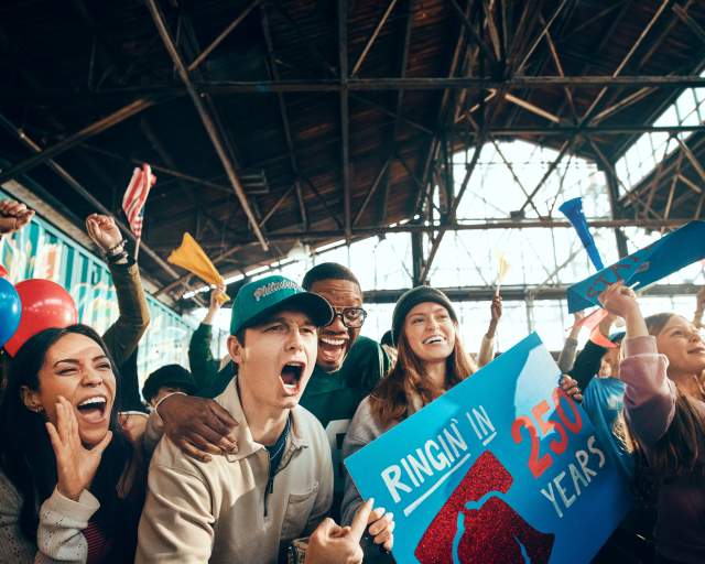 A group of people cheering in a warehouse with a sign that reads Ringin in 250 Years