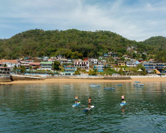 Three people practice paddleboarding in calm waters near the beach and colorful houses on the hillside of Taboga Island, surrounded by lush vegetation.