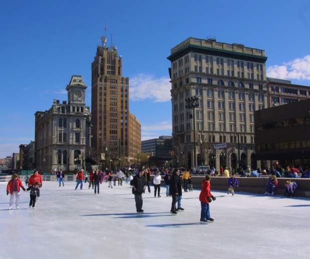 People skate on the outdoor ice rink in Clinton Square on a sunny winter day, with historic downtown Syracuse buildings rising in the background and groups of visitors gathered along the edges of the rink.