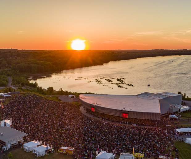 Aerial shot of Lakeview Amphitheater, large crowd infront of amphitheater set on the shores of Onondaga Lake, a fiery orange sunset can be seen in the horizon