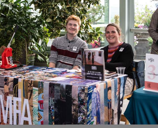 Vendor table covered in a colorful cloth that says UMFA, has swag on it and 2 smiling people sitting behind it.