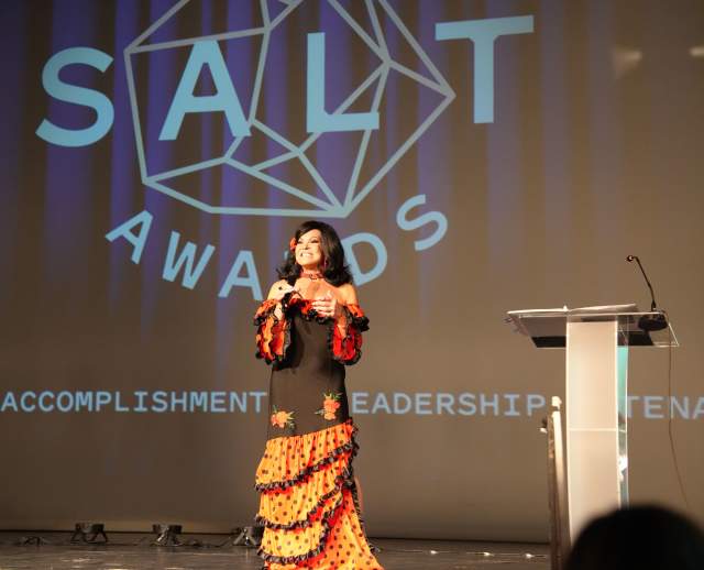 Backdrop says Salt Awards in big text with latina woman in brightly colored dress with a microphone speaking