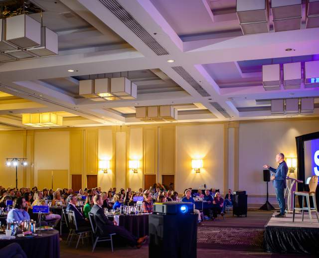 Will Guiderra, a white man in a suit, standing on stage talking to a room full of people sitting at round tables