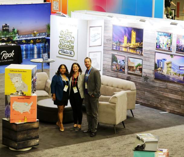 Three smiling team members in a trade show booth surrounded by photos of Sacramento landmarks